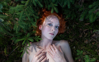 portrait of sexy young redhead girl lies at forest bathing on forest ground between small spruce trees