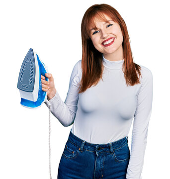 Redhead young woman holding electric steam iron looking positive and happy standing and smiling with a confident smile showing teeth
