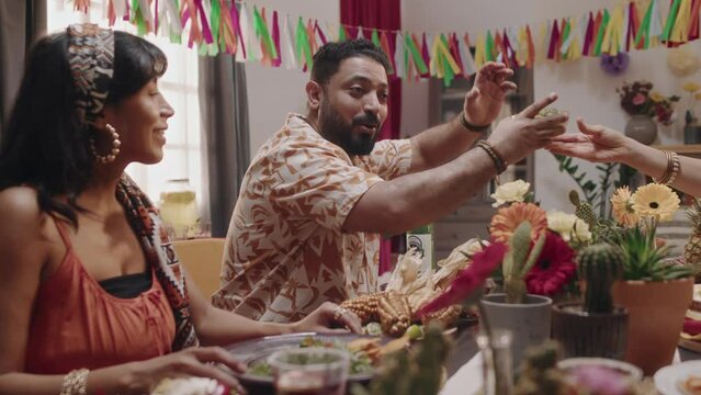 Medium shot of cheerful bearded Mexican man in patterned shirt sitting at dinner table with friends during Cinco de Mayo celebration, telling funny story, wife listening and laughing