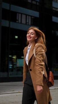 Vertical view of young happy businesswoman in earphones walking the city street while listening music on smartphone, funny dancing