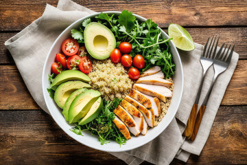 Healthy salad bowl with quinoa, tomatoes, chicken, avocado, lime and mixed vegetables, lettuce, parsley on wooden background. Food and health.
