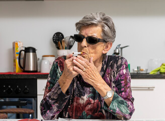 Elderly lady with dark glasses, lights a cigarette in the kitchen in front of her newspaper. - 774759542