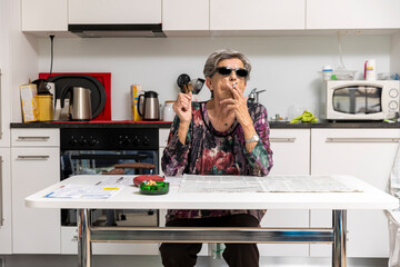 Old lady full of wrinkles smokes comfortably at home in her kitchen wearing black sunglasses. Funny situation - 774759572