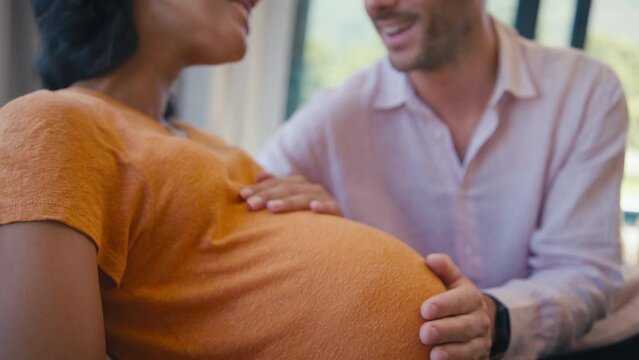 Loving multi-racial couple with pregnant woman and man touching her stomach sitting on sofa at home - shot in slow motion