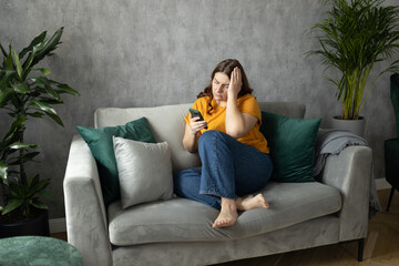 Portrait of beautiful young woman with depressed facial expression sitting on the couch holding her...