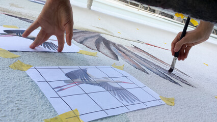 female hands paint a mural of a stork on a scaffolding on a skyscraper according to a gridded template