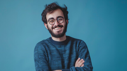 A smiling man with glasses and arms crossed wearing a casual blue sweater against a blue background. - obrazy, fototapety, plakaty