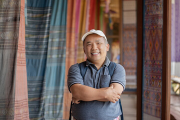 Asian middle-aged man in white cap and t-shirt standing and crossing one's arm in woven fabric shop...