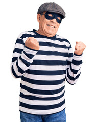 Senior handsome man wearing burglar mask and t-shirt very happy and excited doing winner gesture...