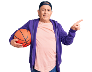 Senior handsome grey-haired man holding basketball ball smiling happy pointing with hand and finger to the side