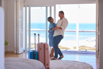 Mature Couple With Luggage Dancing In Beachfront House Overlooking Ocean For Summer Vacation