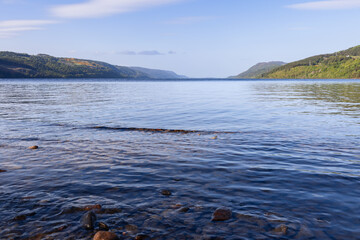 A serene view of Loch Ness under a clear sky, showcasing its vast expanse of water bordered by lush, green hills that gently slope down to meet the shoreline