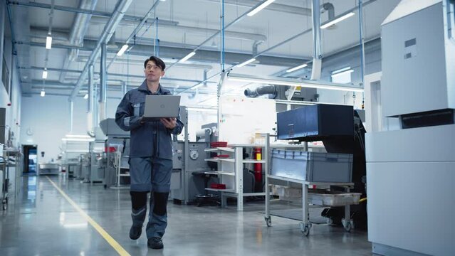 Electronics Production Factory: Confident Asian Male Walking Next to Manufacturing Machines and Using Laptop Computer. Industrial Engineer Monitoring Workflow, Carrying Out Quality Control Operations