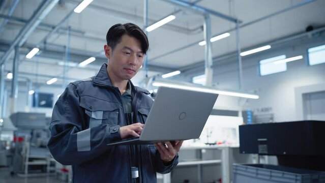 Portrait of a Professional Heavy Industry Asian Engineer Worker in Grey Uniform Working on a Laptop Computer on an Assembly Plant. Confident Industrial Specialist Standing in a Factory Facility