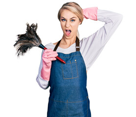 Beautiful young blonde woman wearing apron holding cleaning duster crazy and scared with hands on head, afraid and surprised of shock with open mouth