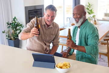Two Mature Male Friends At Home Drinking Beers And Watching Sports On Digital Tablet