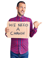 Young handsome man holding we need a change banner pointing thumb up to the side smiling happy with open mouth