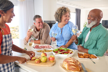 Group Of Smiling Mature Friends At Home Relaxing Preparing Food For Lunch With Wine Together