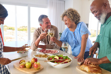 Group Of Smiling Mature Friends At Home Relaxing Preparing Food For Lunch With Wine Together