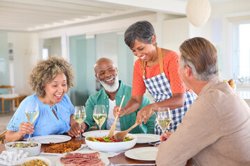 Group Of Smiling Mature Friends At Home Relaxing Meeting For Lunch And Wine Together As Woman Serves