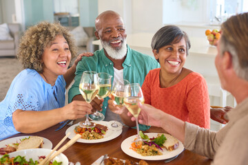 Group Of Smiling Mature Friends At Home Sitting Around Table Meeting For Lunch And Wine Together 