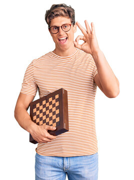Young handsome man holding chess doing ok sign with fingers, smiling friendly gesturing excellent symbol