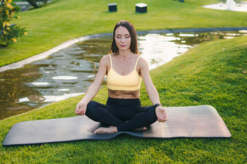 Sporty brunette woman in sportswear sitting on black fitness mat with closed eyes in lotus pose, practicing yoga at public park. Yoga at park. Concept of