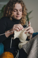 Young woman wearing warm sweater resting on the armchair at home holding knitting needles. Young woman knitting handmade at home.