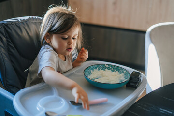 Portrait of cute adorable caucasian child kid girl sitting in high chair eating rice porridge with spoon. Everyday lifestyle. Candid real authentic moment