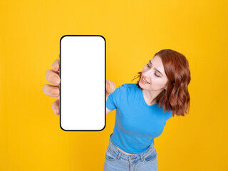 Digital mobile application offer, young caucasian red hair woman showing looking empty blank white screen phone mock up close up to camera. Yellow studio background. Advertisement concept idea image. - Powered by Adobe