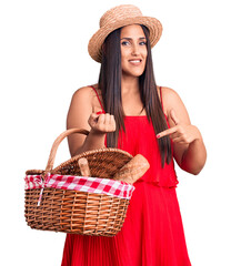 Young beautiful brunette woman wearing summer hat holding picnic basket smiling happy pointing with hand and finger