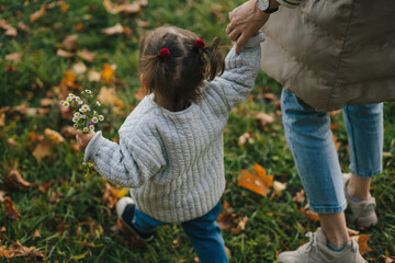 Toddler girl having fun outdoors on autumn day, exploring nature holding mother's hand in a city park. Autumn activities for small kids.