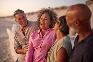 Mature Couple With Friends Sitting Outdoors On Beach Watching Sunset Together