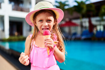 Happy girl in pink swimsuit and sunhat enjoys strawberry ice cream by poolside. Child indulges in frozen treat, summer vibes. Blue water background, leisure day at resort. Fashionable kid swimwear.