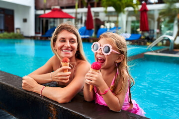 Smiling mom and daughter enjoy ice cream by poolside. Girl in pink swimsuit, flower sunglasses has...