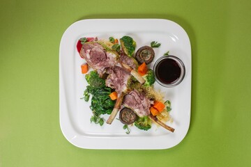 Baked lamb with vegetables on a white platter with red sauce top view on a green background