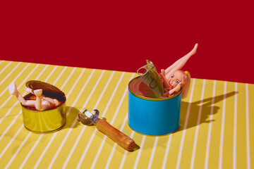 Doll parts in tin cans with can opener on striped tablecloth against red background. Creative blog content about unique food presentation. Concept of pop art photography, creativity