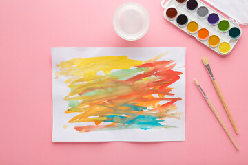 Paintbrushes, watercolor palette and drawn colorful scratches on white paper. Light pink table background. Pastel color. Closeup. Baby development. Learning painting. Top down view.
