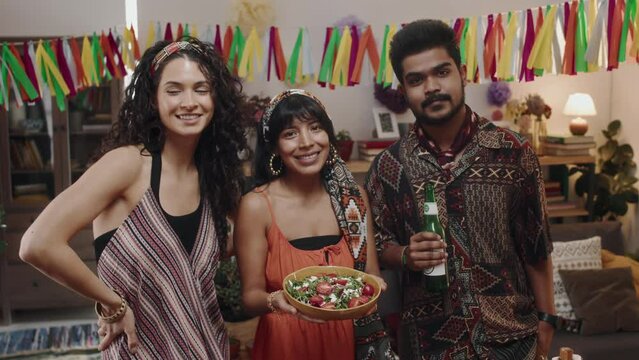 Medium close-up portrait shot of three Mexican people, 2 girls in colourful dresses, holding bowl of salad, and bearded man with beer, posing for fun photos under ribbon garlands at house party