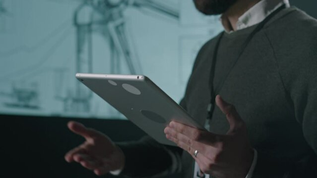 Side tilt footage of male Biracial wind power engineer in glasses and with tablet giving presentation of advancements in windmill production industry to audience during meeting indoors