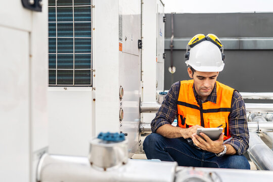 Male technical foreman in safety uniform inspects maintenance work holding a tablet to look at plumbing and electrical systems on the roof of a building, Technician man worker checking power system