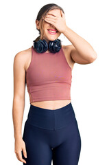 Beautiful caucasian young woman wearing gym clothes and using headphones smiling and laughing with hand on face covering eyes for surprise. blind concept.