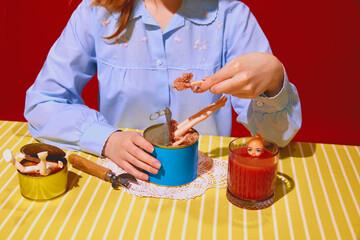 Young woman in blue blouse with doll parts on meat can, drinking tomato juice against red background. Psychological effects of food on mood. Concept of pop art photography, creativity, surrealism