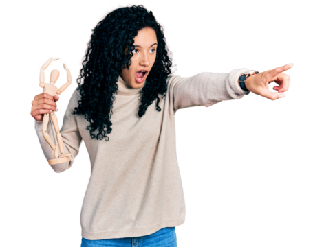 Young hispanic woman with curly hair holding small wooden manikin pointing with finger surprised ahead, open mouth amazed expression, something on the front