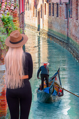 A stylish blonde woman on the bridge watching the Venetian gondolier - Venetian gondolier punting gondola through green canal waters of Venice Italy - obrazy, fototapety, plakaty