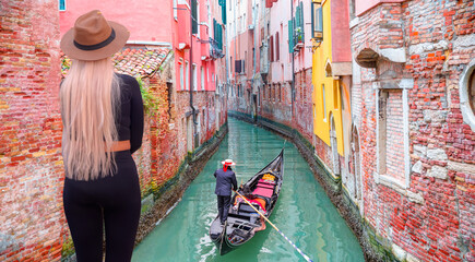 A stylish blonde woman on the bridge watching the Venetian gondolier - Venetian gondolier punting gondola through green canal waters of Venice Italy - obrazy, fototapety, plakaty