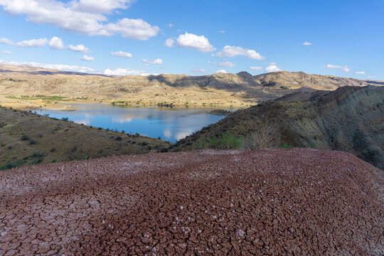 Beautiful view of the lake and mountains. Red earth in the foreground. Bright blue sky. Clouds are reflected in the water. Dry grass. Mravaltskaro Reservoir in Georgia.
