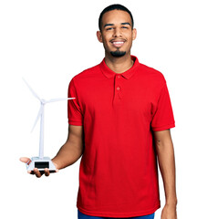 Young african american man holding solar windmill for renewable electricity looking positive and...