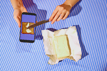 Hands buttering virtual toast on a smartphone screen with real butter and knife. Ad for a cooking app showcasing interactive and digital recipe tutorials. Concept of pop art photography, creativity