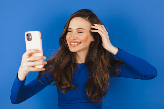Brunette girl stares at the phone and fixes her hair. Female uses phone like mirror. Young girl make photo from hands with phone on blue background.